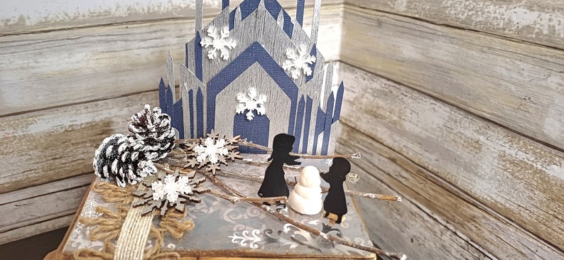 May include: A miniature winter scene with a blue and gray paper castle, two black paper figures, a white paper snowman, and brown pine cones on a wooden surface.
