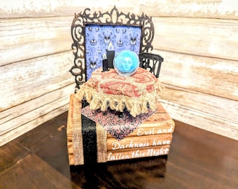 May include: A miniature Halloween display with a small black chair, a decorative frame with a blue background, and a crystal ball. The display sits on a stack of books with the text "Evil and Darkness have fallen this Night."
