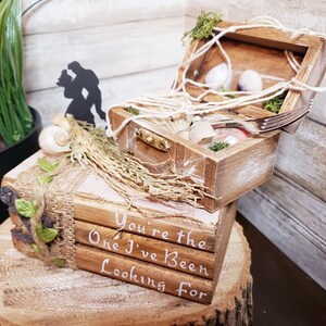 K&ouml;nnte beinhalten: Eine Holzkiste mit Muschel- und Seildesign, einer Silhouette eines Paares und einem Stapel B&uuml;cher mit dem Text "You're the One I've Been Looking For".