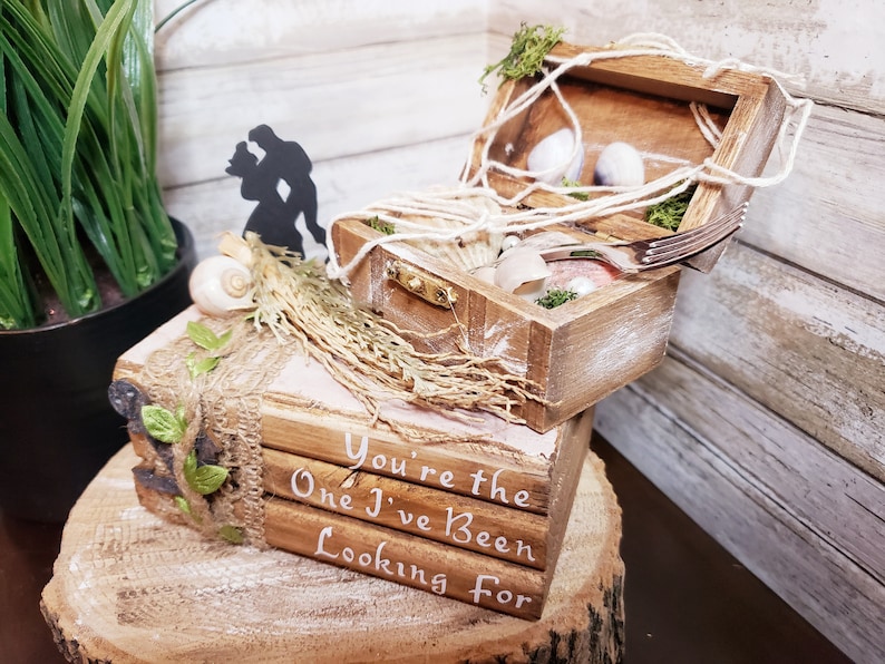 K&ouml;nnte beinhalten: Eine Holzkiste mit Muschel- und Seildesign, einer Silhouette eines Paares und einem Stapel B&uuml;cher mit dem Text "You're the One I've Been Looking For".