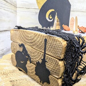 May include: A wooden tiered tray decorated with black paper cutouts of a witch, a broom, and a spooky house. The tray is adorned with black lace and a small black spider. The text "We're simply meant to be" is visible on the tray.