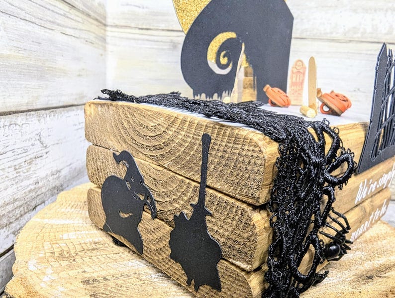 May include: A wooden tiered tray decorated with black paper cutouts of a witch, a broom, and a spooky house. The tray is adorned with black lace and a small black spider. The text "We're simply meant to be" is visible on the tray.