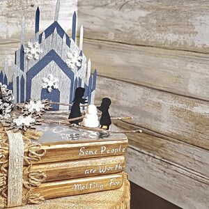 May include: A stack of four books with a winter-themed design. The top book has a blue and white paper castle on top. The books are wrapped in twine and have the text "Some People are Worth Melting For" on the front.