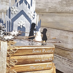 May include: A stack of four books with a rustic brown cover and white text that reads "Some People are Worth Melting For". The books are decorated with a burlap ribbon and a pine cone. A paper cutout of a castle with snowflakes and two figures standing in front of it is on top of the books.