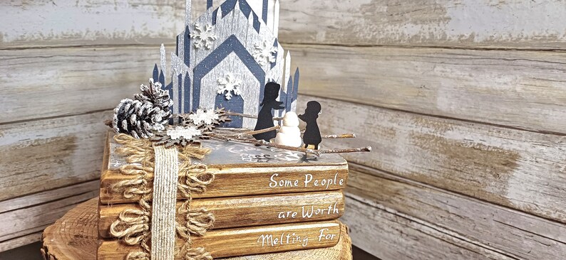 May include: A stack of four books with a rustic brown cover and white text that reads "Some People are Worth Melting For". The books are decorated with a burlap ribbon and a pine cone. A paper cutout of a castle with snowflakes and two figures standing in front of it is on top of the books.