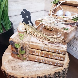 K&ouml;nnte beinhalten: Eine Holzkiste mit einem Seilgriff und einer Silhouette eines Paares obenauf. Die Kiste ist ge&ouml;ffnet und enth&auml;lt Muscheln und Moos. Die Kiste steht auf einem Stapel B&uuml;cher mit dem Text "You're the One I've Been Looking For" darauf.