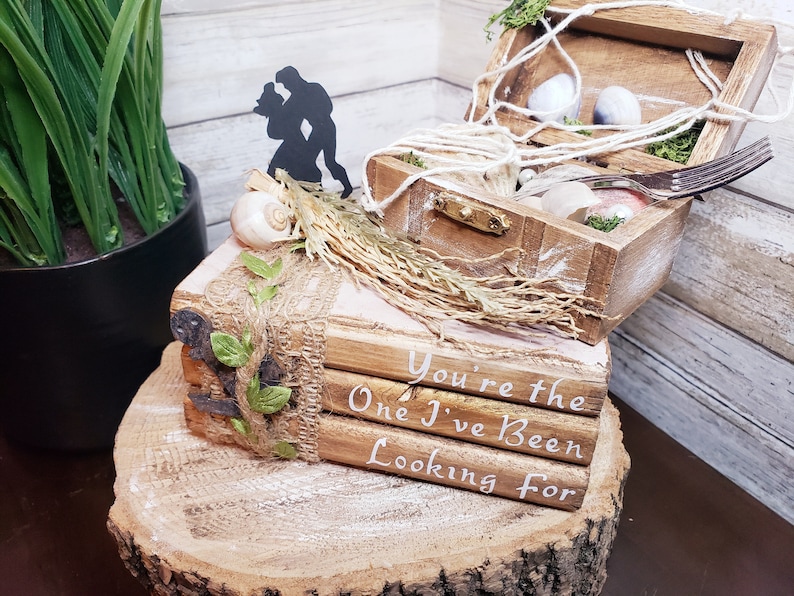 K&ouml;nnte beinhalten: Eine Holzkiste mit einem Seilgriff und einer Silhouette eines Paares obenauf. Die Kiste ist ge&ouml;ffnet und enth&auml;lt Muscheln und Moos. Die Kiste steht auf einem Stapel B&uuml;cher mit dem Text "You're the One I've Been Looking For" darauf.