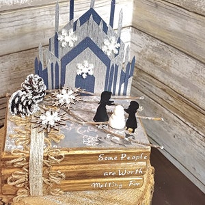 May include: A stack of books decorated with a winter scene. The scene includes a blue and gray castle, two figures, a snowman, and pine cones. The books have a rustic brown cover and the top book has the text "Some People are Worth Melting For".