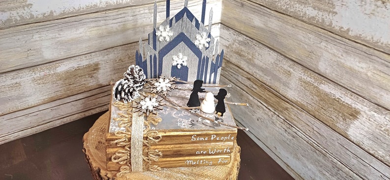 May include: A stack of books decorated with a winter scene. The scene includes a blue and gray castle, two figures, a snowman, and pine cones. The books have a rustic brown cover and the top book has the text "Some People are Worth Melting For".
