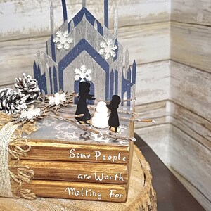 May include: A stack of three books with a winter scene on top. The books are decorated with burlap ribbon and a pine cone. The top book has the text "Some People are Worth Melting For".