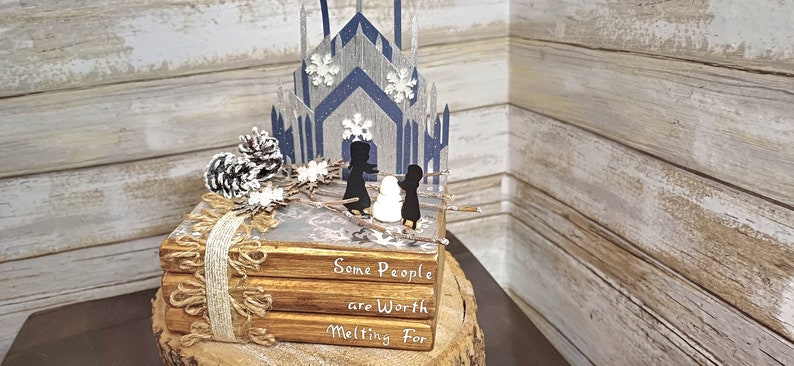 May include: A stack of three books with a winter scene on top. The books are decorated with burlap ribbon and a pine cone. The top book has the text "Some People are Worth Melting For".