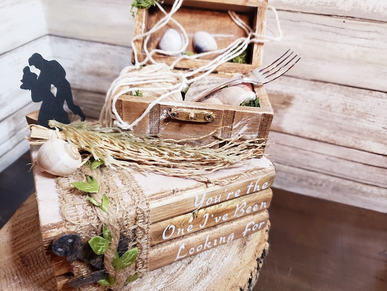 K&ouml;nnte beinhalten: Eine Holzkiste mit rustikalem Design, verziert mit Muscheln, Schnur und einer Gabel. Die Kiste ist auf einer kleineren Holzkiste mit dem Text "You're the One I've Been Looking For" gestapelt.