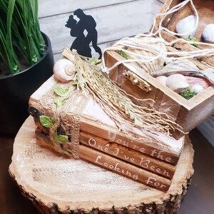 K&ouml;nnte beinhalten: Eine Holzkiste mit einer Muschel und Moos im Inneren, verziert mit einem Juteband und gr&uuml;nen Bl&auml;ttern. Die Kiste steht auf einem Holzsockel mit den Worten "You're the One I've Been Looking For" darauf. Eine Silhouette eines Paares ist oben auf der Kiste angebracht.