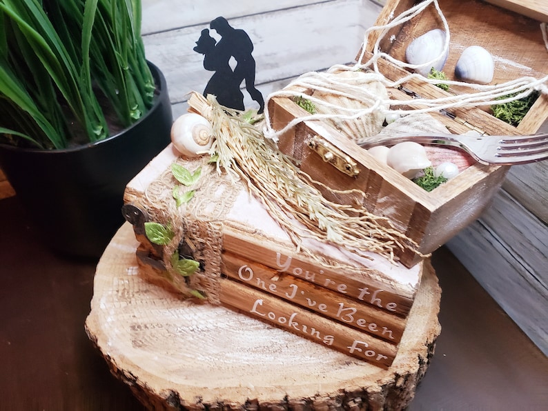 K&ouml;nnte beinhalten: Eine Holzkiste mit einer Muschel und Moos im Inneren, verziert mit einem Juteband und gr&uuml;nen Bl&auml;ttern. Die Kiste steht auf einem Holzsockel mit den Worten "You're the One I've Been Looking For" darauf. Eine Silhouette eines Paares ist oben auf der Kiste angebracht.