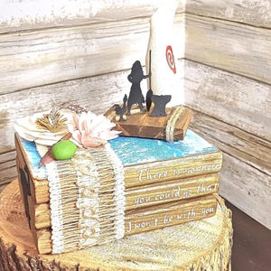 Könnte beinhalten: Ein Stapel von drei Büchern mit einem rustikalen Holzdeckel und einem blau-weiß bemalten Design. Das oberste Buch hat eine Holzsilhouette einer Person, die ein Boot segelt, mit dem Text "Es gibt keinen Ort, an den du gehen könntest, an dem ich nicht bei dir wäre".