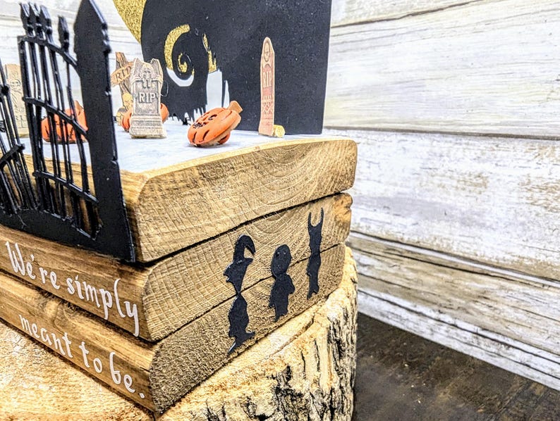 May include: A wooden tiered display with a black silhouette of a gate, a tombstone, and a crescent moon. The display is decorated with the text "We're simply meant to be."