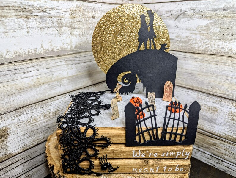 May include: A black and white silhouette of Jack Skellington and Sally from the Nightmare Before Christmas movie, standing on a hilltop with a golden moon behind them. The scene is set in a graveyard with a gate, tombstones, and pumpkins. The text "We're simply meant to be." is written below the image.