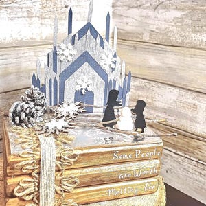 May include: A stack of four books with a winter-themed papercraft scene on top. The scene features a blue and white ice castle, two figures, and a snowman. The books have the text "Some People are Worth Melting For" on the bottom book.