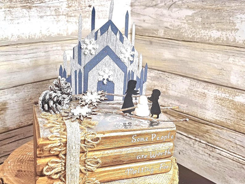 May include: A stack of four books with a winter-themed papercraft scene on top. The scene features a blue and white ice castle, two figures, and a snowman. The books have the text "Some People are Worth Melting For" on the bottom book.
