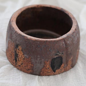 May include: A handcrafted, round ceramic bowl with a rustic, textured appearance. The bowl features a dark brown exterior with areas of lighter, reddish-brown and black accents. The interior is a deep brown. The bowl is approximately 4 inches in diameter and 3 inches tall.