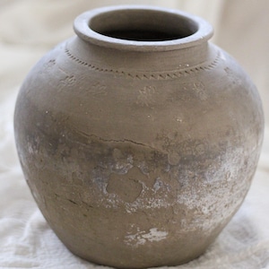 Pode incluir: Um vaso redondo de cerâmica cinza com uma abertura larga e uma superfície texturizada. O vaso tem uma aparência ligeiramente desgastada com manchas brancas e detalhes decorativos perto da borda. Está sobre um tecido branco.