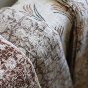May include: A folded textile featuring a layered design with multiple patterns. The top layer displays a cream background with orange and dark green floral motifs, while the lower layers show intricate brown and white botanical prints.
