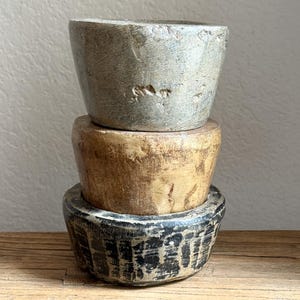 Puede incluir: Tres cuencos de piedra hechos a mano apilados. El cuenco superior es gris, el cuenco del medio es marrón y el cuenco inferior es negro con un patrón texturizado. Los cuencos se exhiben sobre una superficie de madera rústica.