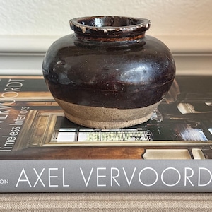 May include: A small, round, earthenware pot with a dark brown glazed upper section and a raw, unglazed beige base. The pot features a wide, short neck with a slightly worn, chipped rim, showcasing a distinct contrast between the smooth, dark glaze and the textured, matte bottom surface.