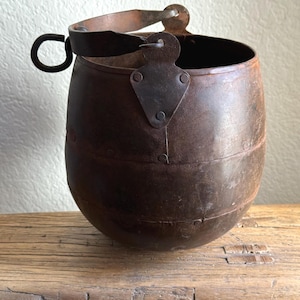 May include: A rustic, antique metal bucket with a rounded base and a handle. The bucket is a dark brown color, showing signs of age and wear. It has a metal handle and a hinged bracket for the handle. The bucket is sitting on a wooden surface.