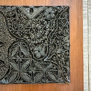 Könnte beinhalten: Eine dekorative Fliese mit schwarzem Hintergrund und weißem Blumenmuster, eingefasst in einen Holzrahmen. Das komplizierte Design zeigt Blumen, Blätter und geometrische Formen und erzeugt einen detaillierten und künstlerischen Look.