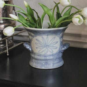 Könnte beinhalten: Eine Keramikvase mit einem hellblauen und weißen handgemalten Blumenmuster, ausgestattet mit zwei seitlichen Griffen und einem breiten Rand. Die Vase ist mit mehreren weißen Tulpen mit grünen Stielen und Blättern gefüllt und hat eine gesprenkelte Glasur.