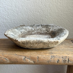 May include: A shallow, oval-shaped stone bowl with a rough, weathered texture and mottled gray and beige coloring. The basin features an uneven, natural rim and a concave center, showcasing a porous, aged surface with visible mineral variations.