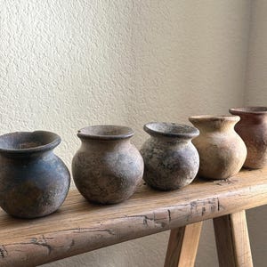 May include: Five small, aged ceramic pots with rounded bodies and wide openings. The pots are in varying shades of brown, grey, and blue, and sit on a rustic wooden bench. The background is a textured, off-white wall.