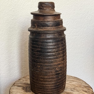May include: A dark brown cylindrical wooden container with a ribbed texture along the body. The vessel features a tiered, stepped lid with a small knob at the top. The wood displays a weathered, deep brown finish with visible grain and horizontal lines.