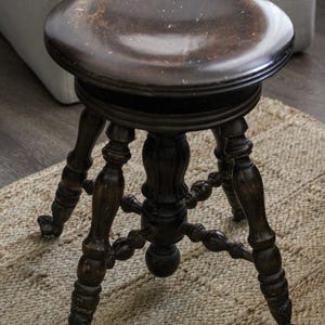 May include: A dark brown wooden stool with a circular seat and four ornate, turned legs. The legs feature decorative carvings and terminate in small feet. A central support column connects the legs, providing structural stability to the piece.