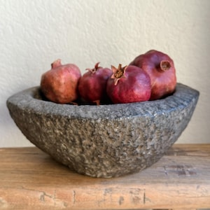 Könnte beinhalten: Eine rustikale, graue Steinschale gefüllt mit mehreren reifen Granatäpfeln. Die Granatäpfel haben eine tiefrote Farbe, eine strukturierte Schale und sichtbare Kronen. Die Schale steht auf einer verwitterten Holzoberfläche und erzeugt eine natürliche, erdige Ästhetik.