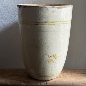 May include: A tall, off-white ceramic vase with a slightly tapered shape. The vase features two horizontal yellow bands and a faded floral design. It has a rustic, aged appearance, suggesting it is vintage or antique.