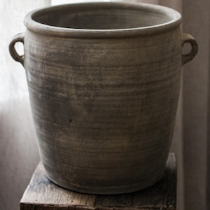 May include: A large, weathered gray ceramic planter with two small handles. The pot has a wide opening and sits on a rustic wooden block. The neutral tones and simple design suggest a minimalist aesthetic.