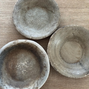 Könnte beinhalten: Drei graue Steinschalen mit rustikalem, verwittertem Aussehen. Die Schalen haben eine flache, abgerundete Form und unterschiedliche Grautöne mit dunkleren Rändern. Die Schalen sind auf einer hellbraunen Holzoberfläche angeordnet.