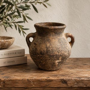 May include: A rustic, weathered brown clay vessel with a wide mouth and two side handles. The surface features a mottled, dark-toned texture with visible signs of age and wear, showcasing a rounded body and a thick, flared rim design.