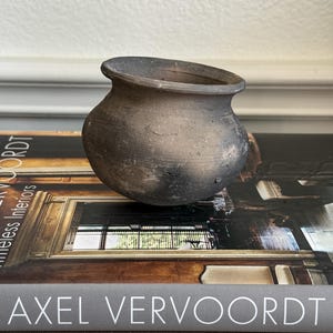 May include: A small, round, dark gray ceramic pot with a wide, flared rim and a textured, matte finish. The pot has a bulbous body and sits on a book titled Axel Vervoordt, which features a photograph of an interior space on its cover.