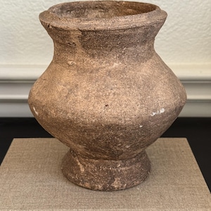 May include: A small, weathered, brown earthenware vessel with a wide, rounded body, a flared rim, and a pedestal base. The surface of the pot has a rough, matte texture with visible signs of age, including minor chips and uneven coloration.