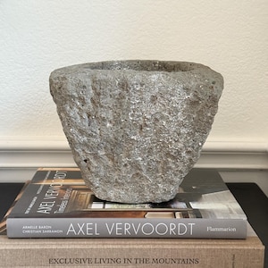 Könnte beinhalten: Ein graues Steingefäß mit rauer Textur und breiter Öffnung. Das Gefäß steht auf zwei Büchern mit dem Titel "Axel Vervoordt". Die Bücher sind auf einer dunklen Oberfläche vor einer weißen Wand gestapelt.