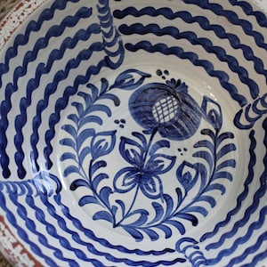 May include: A white ceramic bowl with a blue wave pattern around the rim and a floral design in the center. The bowl has a distressed, rustic look with a red-brown rim.