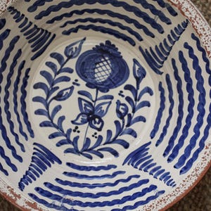 Può includere: Una ciotola rotonda in ceramica con interno color crema caratterizzata da un motivo floreale blu dipinto a mano al centro. Le pareti interne sono decorate con linee blu ondulate concentriche. Il bordo presenta una finitura bianca.