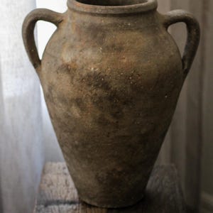 May include: A rustic, brown ceramic vase with two loop handles. The vase has a wide body that tapers to a narrow base. It sits on a wooden block. The vase has a rough, textured surface.