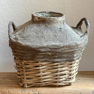 May include: A handcrafted decorative vase with a woven basket base and a textured, gray paper-like top. The vase has two curved handles and a wide opening. The neutral tones and rustic design create a vintage aesthetic.