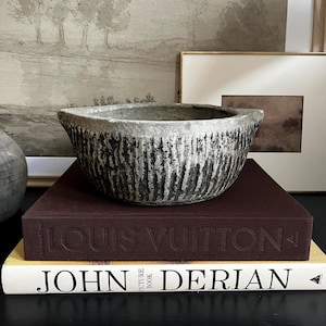 May include: A grey concrete bowl with a textured surface sits on top of two books. The top book is a dark burgundy colour with the text "LOUIS VUITTON" embossed on the cover. The bottom book is white with the text "JOHN DERIAN PICTURE BOOK" printed on the cover.