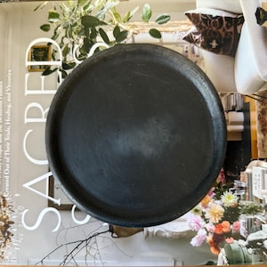 Puede incluir: Una bandeja redonda de color gris oscuro con un borde ligeramente elevado. La bandeja es de un material mate y parece estar vacía. La bandeja está colocada sobre una revista con la palabra "SACRED" visible.