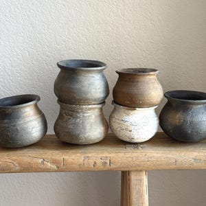 May include: A collection of small, rustic ceramic pots in various shades of gray, brown, and white. The pots are arranged on a weathered wooden surface, showcasing their unique textures and shapes. The pots have a rounded body and a slightly flared rim.